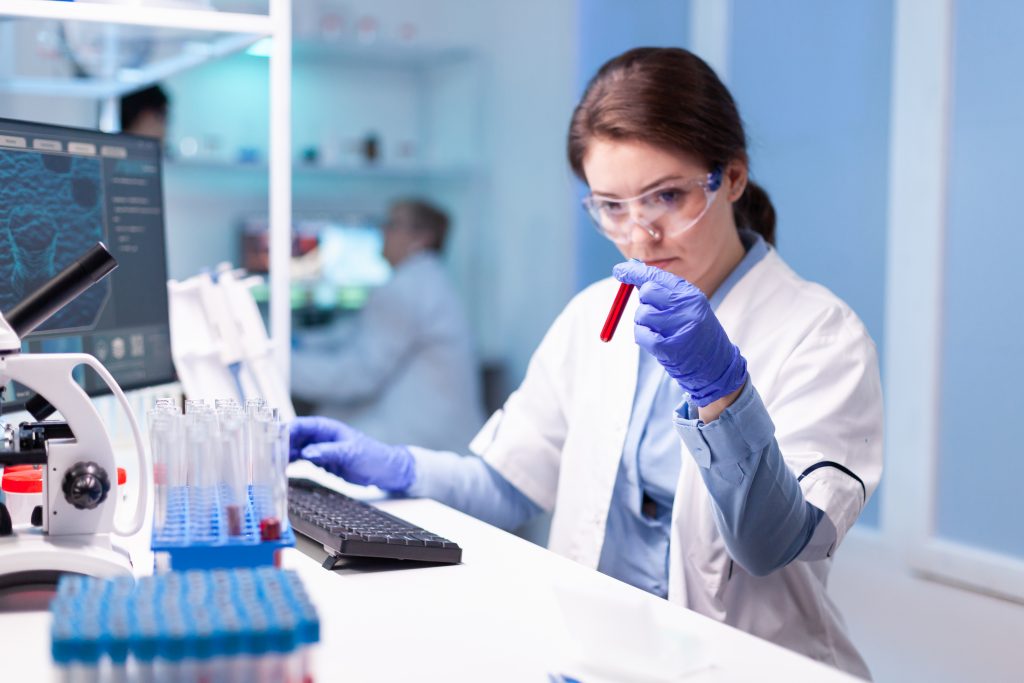 Laboratorium badawcze. Przy stanowisku laboratoryjnym osoba w okularach ochronnych analizuje próbkę w probówce. Na stole znajdują się probówki, mikroskop oraz komputer wykorzystywane w badaniach laboratoryjnych.