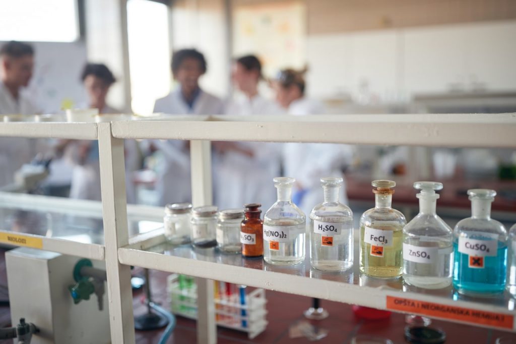 Laboratorium chemiczne. Na pierwszym planie znajduje się rząd butelek z odczynnikami chemicznymi ustawionych na półce laboratoryjnej, a w tle widoczne są osoby pracujące w laboratorium.